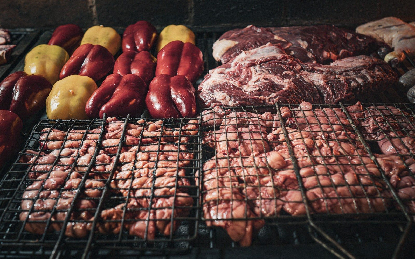 Raw meats and peppers laid out on wire grates over a charcoal grill