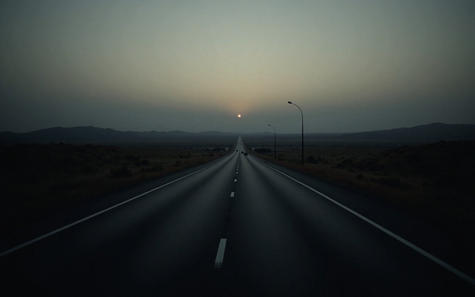 A hazy sun sets over a straight desert highway flanked by distant mountains
